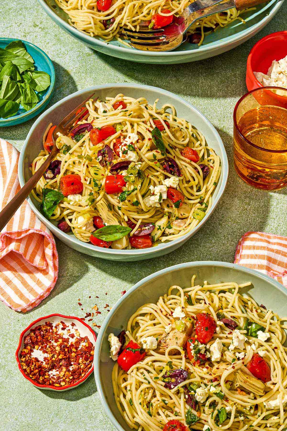 A serving of Mediterranean pasta in a bowl with a fork. Next to it is another bowl of pasta, a glass of sparkling water and bowls of fresh basil, feta and red pepper flakes.