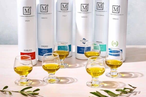 The Mediterranean Dish olive oil bottles lined up on a table with 6 olive oil tasting glasses.