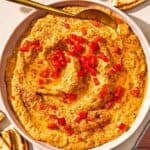 Roasted red pepper hummus in a bowl with a spoon. Nearby - a bottle of olive oil and a few pieces of pita bread.