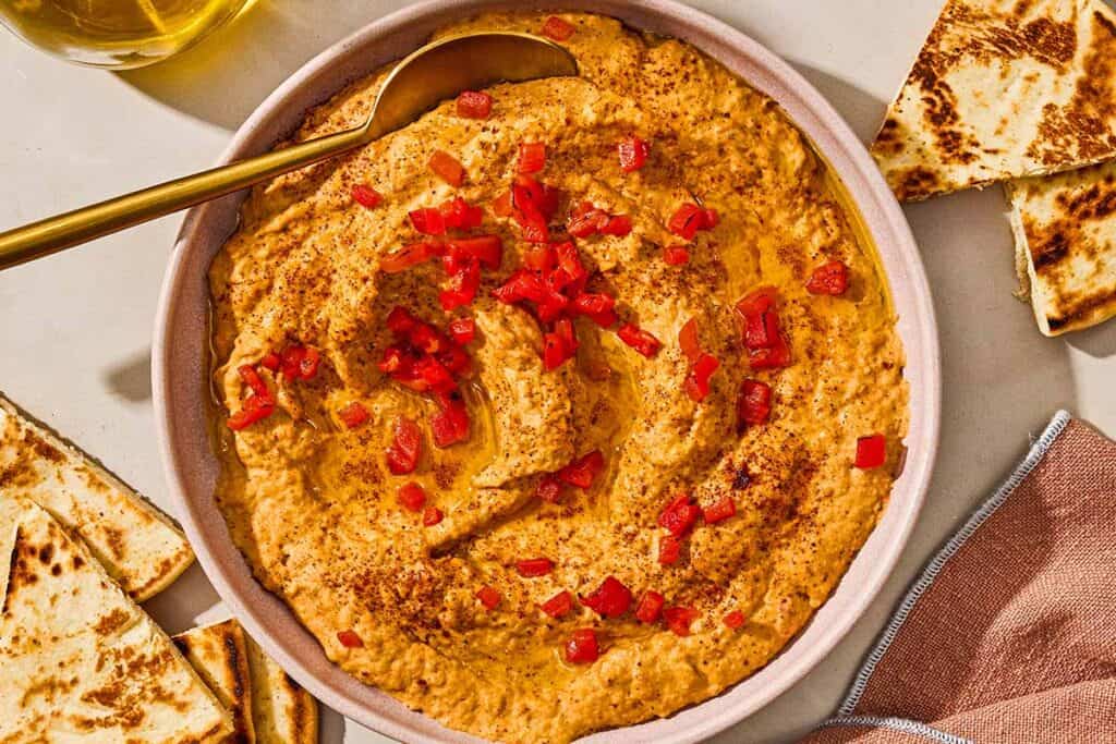 Roasted red pepper hummus in a bowl with a spoon surrounded by several pita wedges.