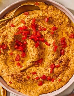 Roasted red pepper hummus in a bowl with a spoon surrounded by several pita wedges.