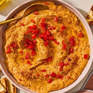 Roasted red pepper hummus in a bowl with a spoon surrounded by several pita wedges.