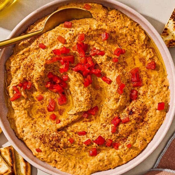 Roasted red pepper hummus in a bowl with a spoon surrounded by several pita wedges.