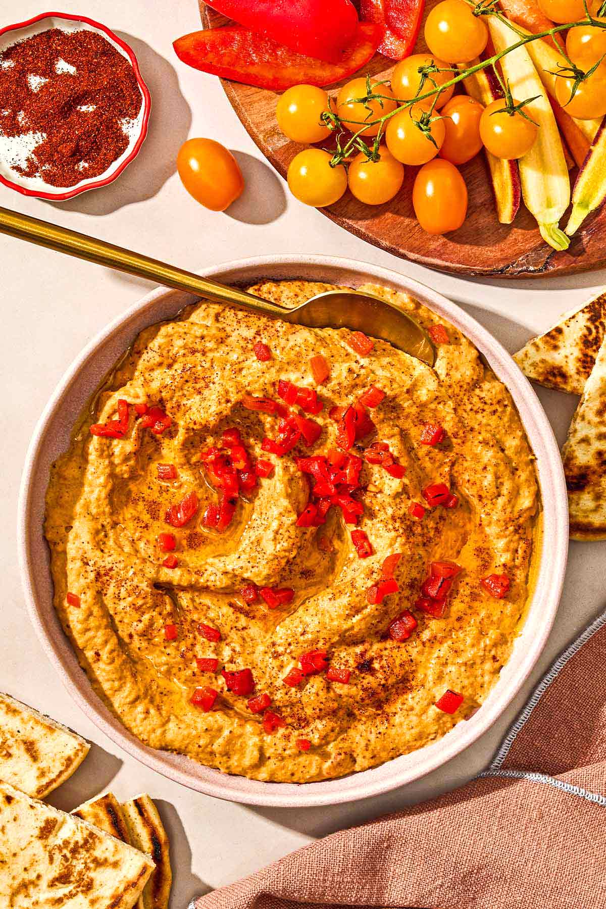 Roasted red pepper hummus in a bowl with a spoon. Nearby is a bowl of sumac, a plate of vegetables and a few pieces of pita bread.