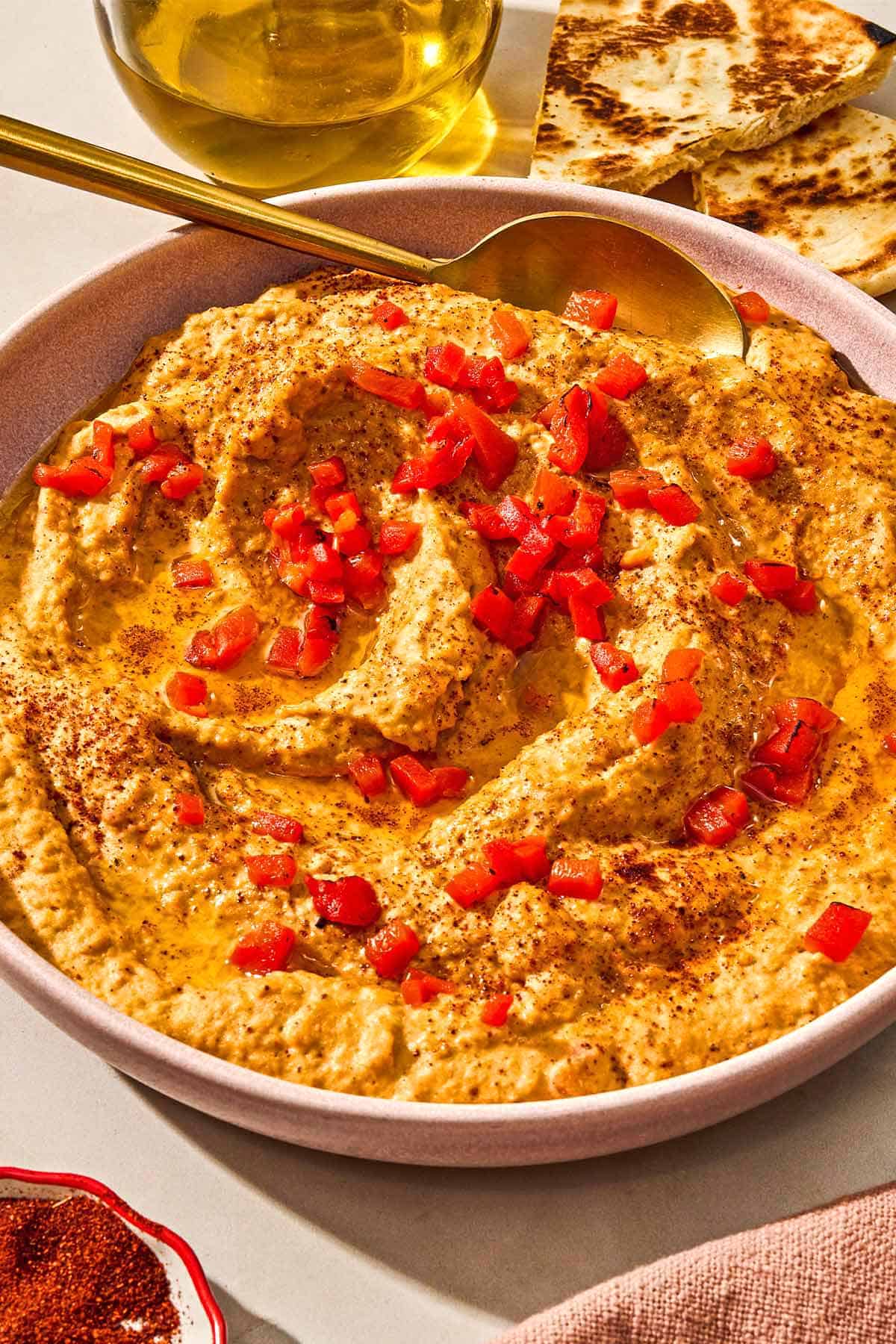 Roasted red pepper hummus in a bowl with a spoon. Next to this is a bowl of sumac, a plate of veggies and several pita wedges.