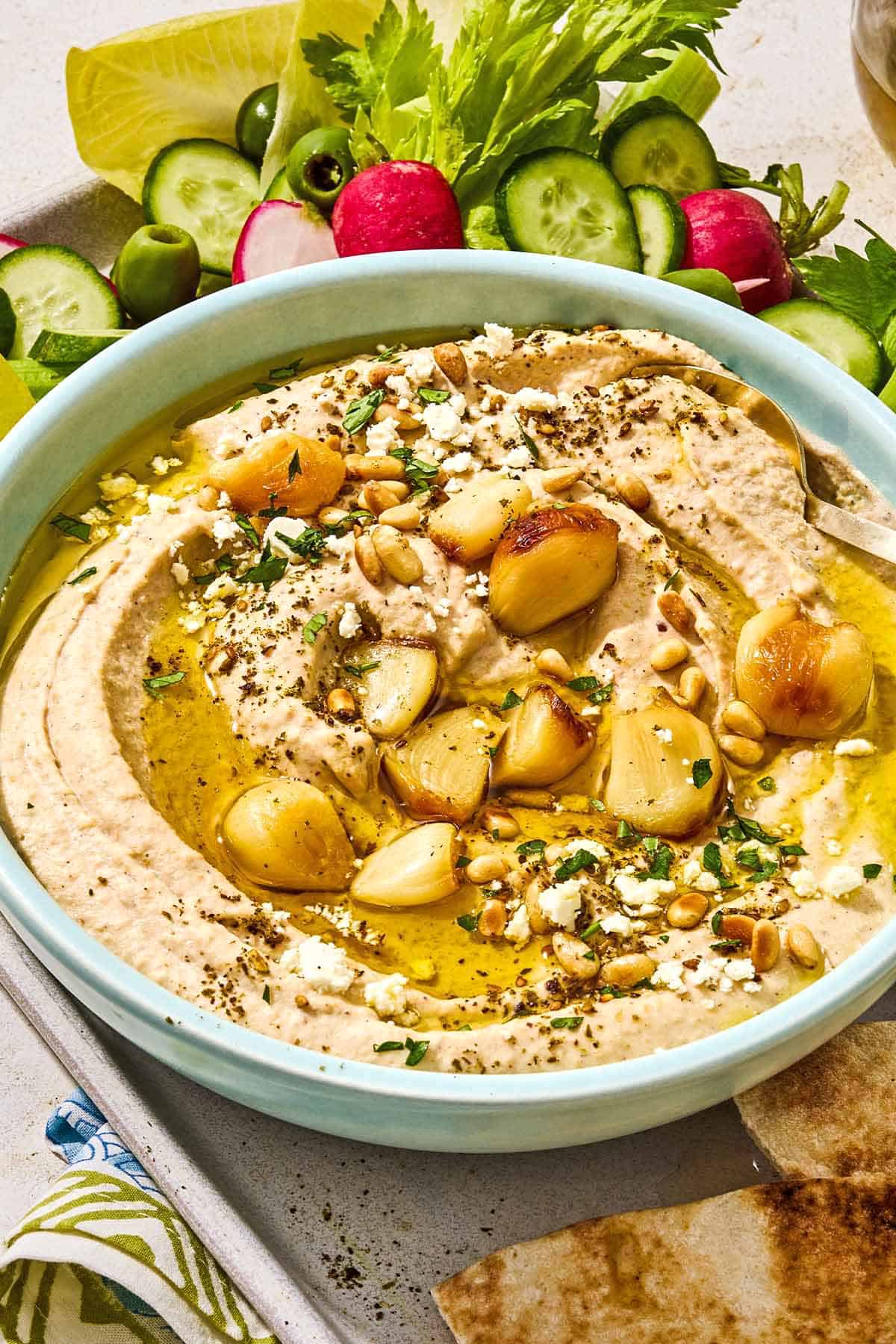 Roasted garlic hummus in a bowl on a tray with sliced vegetables, pita bread wedges and a bowl of crumbled feta.