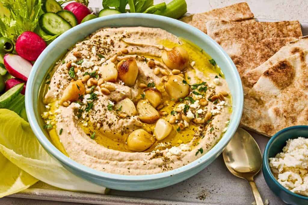 Roasted garlic hummus in a bowl on a tray with sliced vegetables, pita bread wedges and a bowl of crumbled feta.