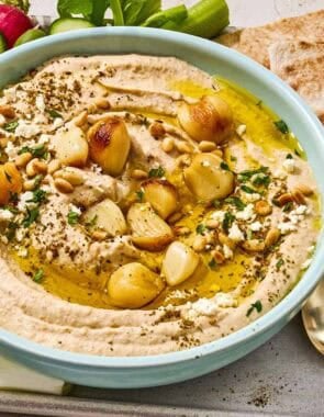 Roasted garlic hummus in a bowl on a tray with sliced vegetables, pita bread wedges and a bowl of crumbled feta.