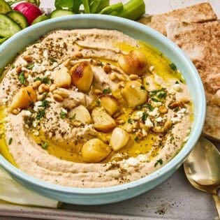 Roasted garlic hummus in a bowl on a tray with sliced vegetables, pita bread wedges and a bowl of crumbled feta.