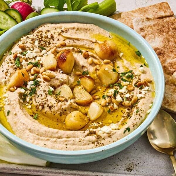 Roasted garlic hummus in a bowl on a tray with sliced vegetables, pita bread wedges and a bowl of crumbled feta.