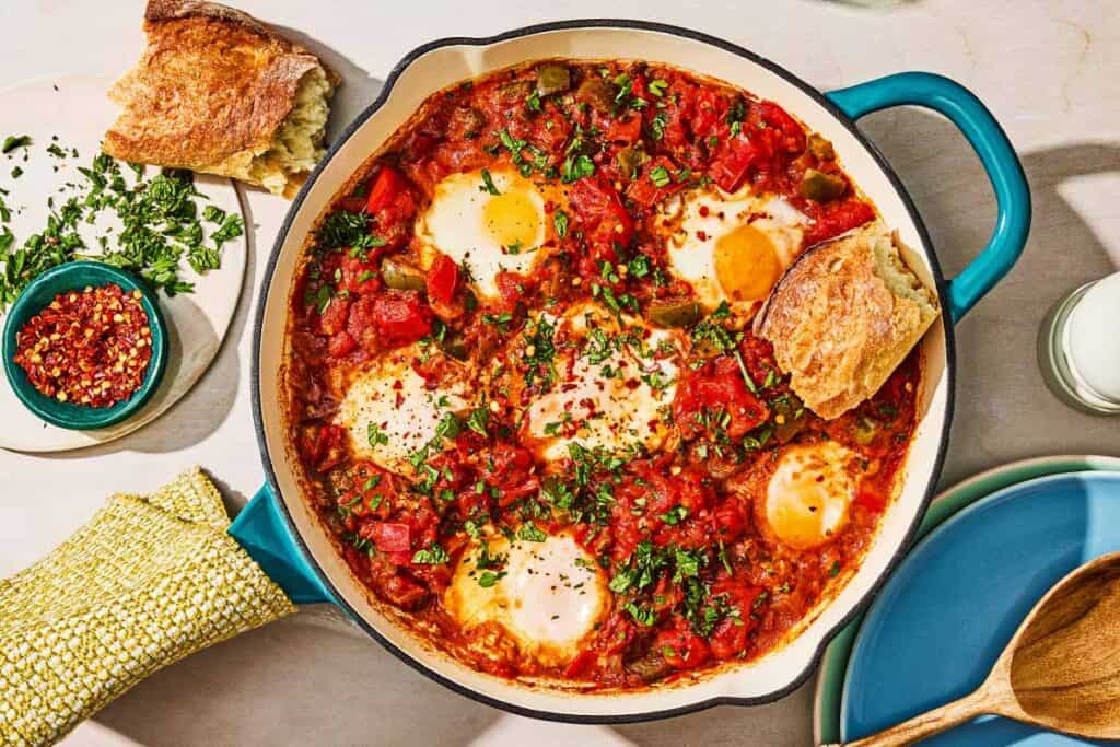 The shakshuka recipe in a skillet next to a bowl of red pepper flakes, a piece of crusty bread, and a stack of plates with a wooden spoon.