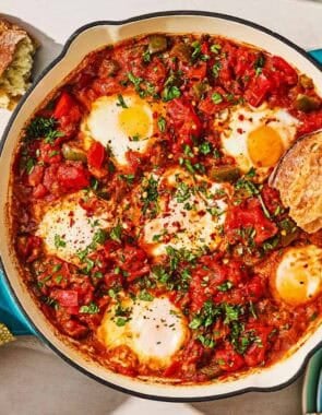 The shakshuka recipe in a skillet next to a bowl of red pepper flakes, a piece of crusty bread, and a stack of plates with a wooden spoon.