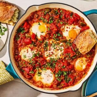 The shakshuka recipe in a skillet next to a bowl of red pepper flakes, a piece of crusty bread, and a stack of plates with a wooden spoon.