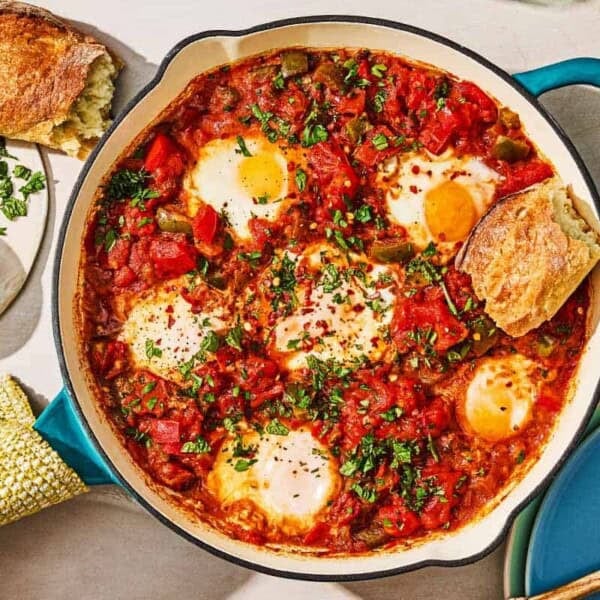 The shakshuka recipe in a skillet next to a bowl of red pepper flakes, a piece of crusty bread, and a stack of plates with a wooden spoon.