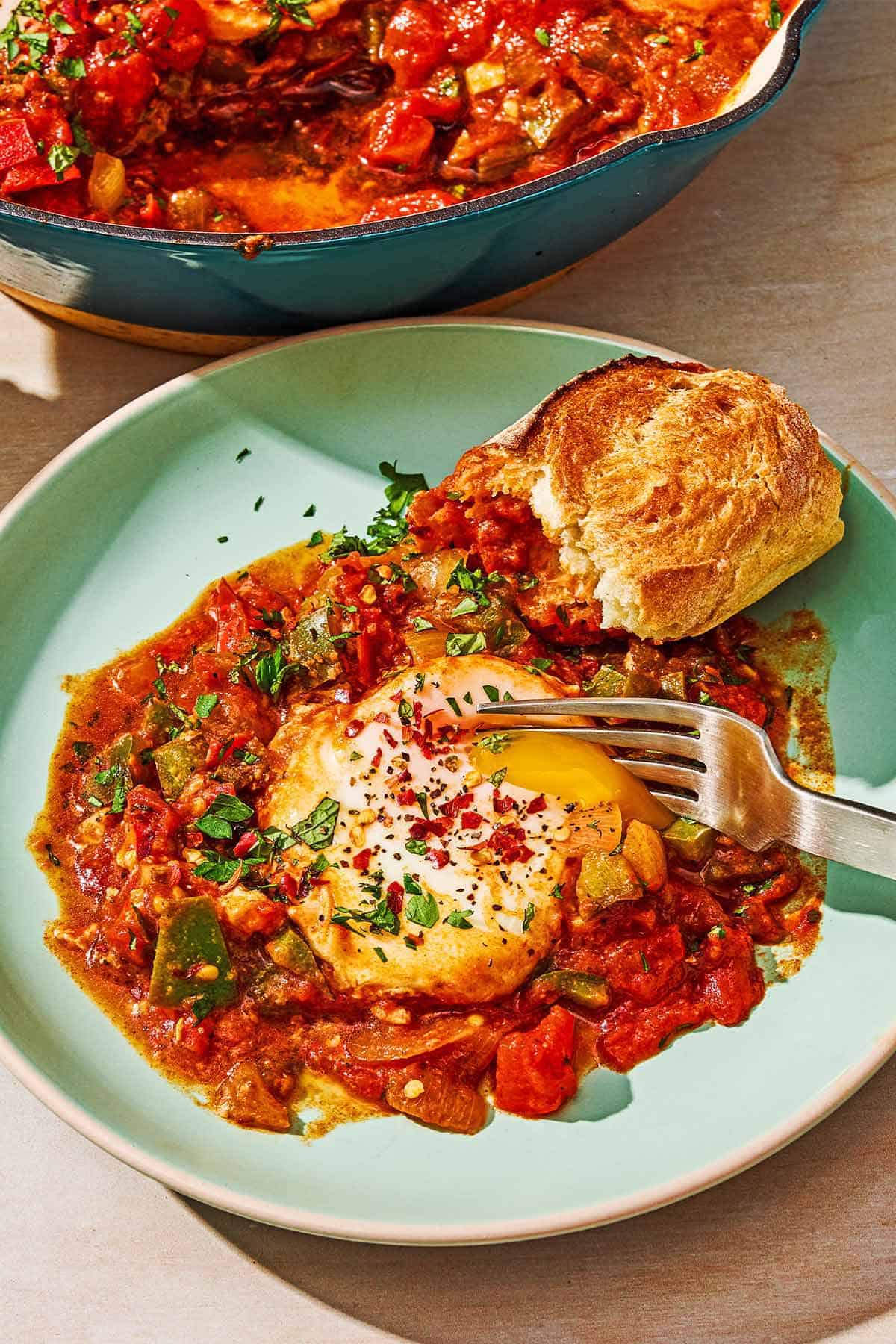 A serving of the shakshuka recipe on a plate with a piece of crusty bread and a fork. Next to this is the rest of the shakshuka in a skillet.