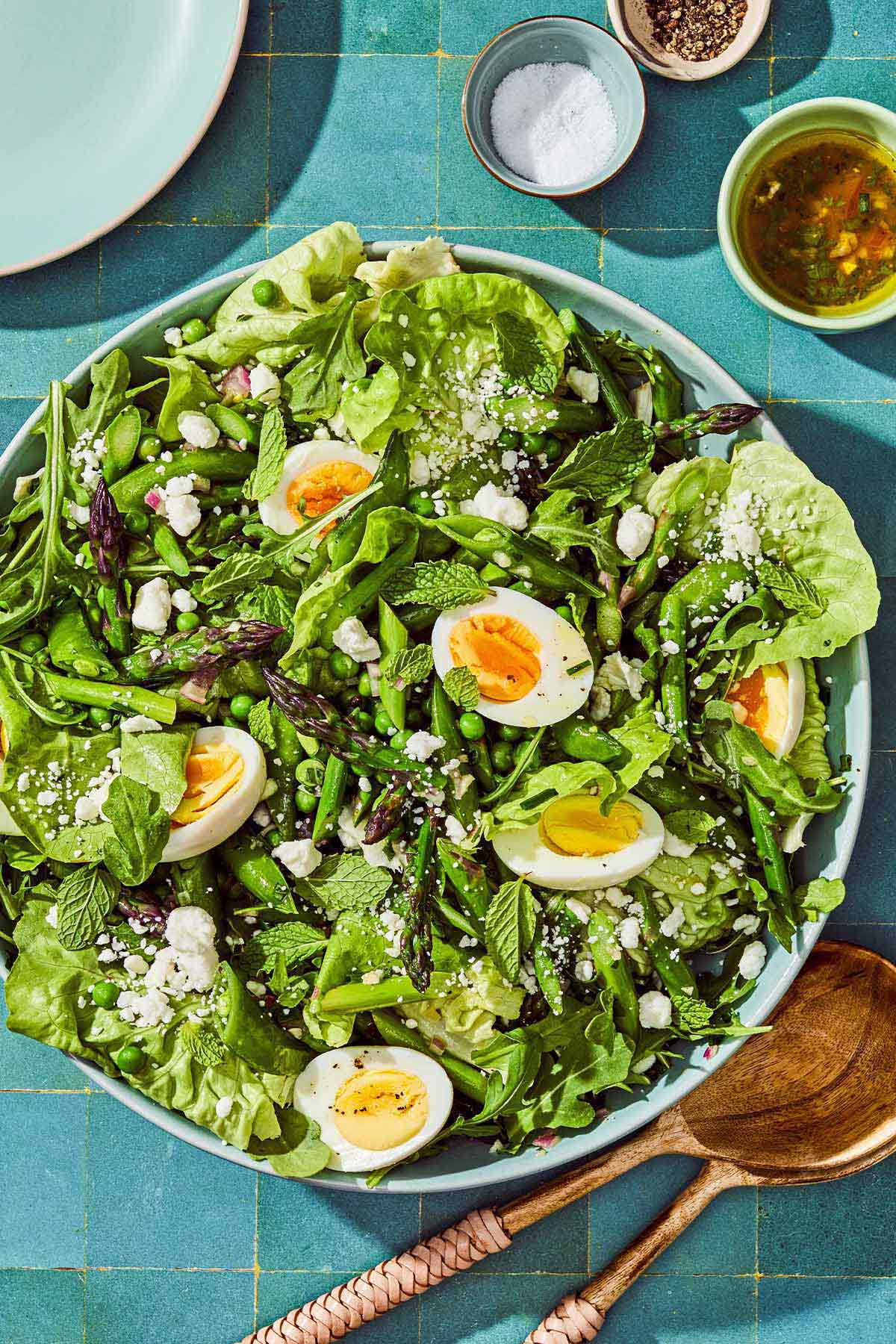 A spring green salad in a serving bowl next to serving utensils, bowls of salt and pepper, a bowl of the dressing, and a plate.
