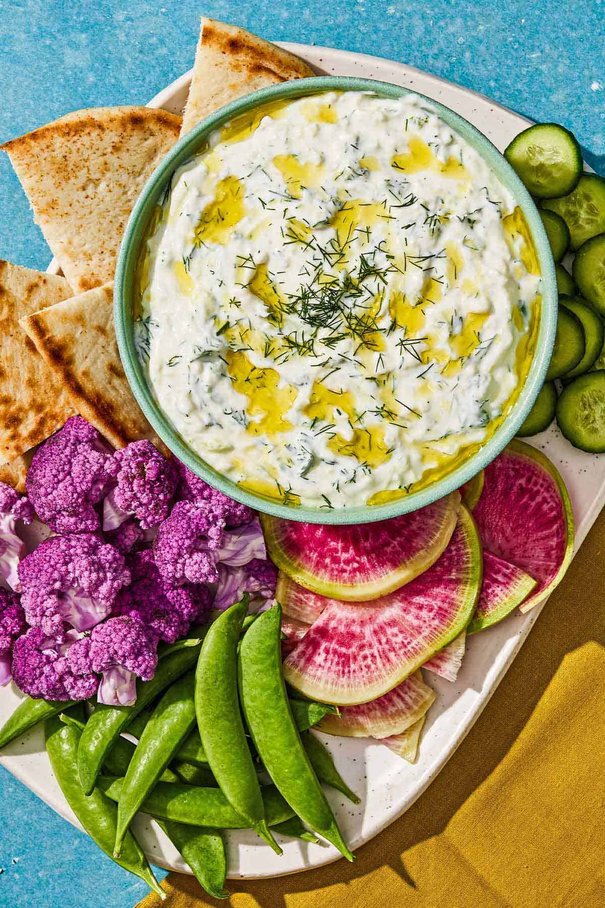 Tzatziki in a bowl with a spoon on a platter with vegetables and pita wedges.