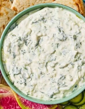 Tzatziki in a bowl with a spoon on a platter with vegetables and pita wedges.
