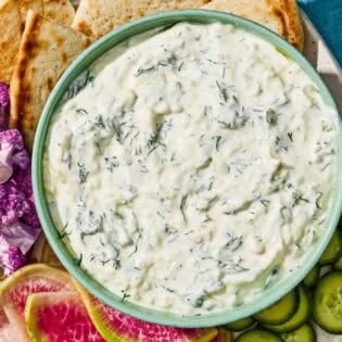 Tzatziki in a bowl with a spoon on a platter with vegetables and pita wedges.