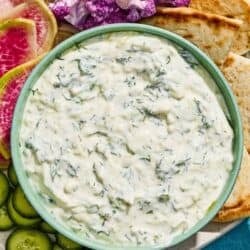 Tzatziki in a bowl with a spoon on a platter with vegetables and pita wedges.