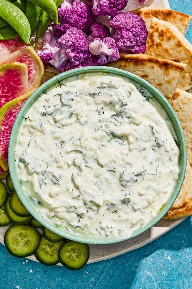 Tzatziki in a bowl with a spoon on a platter with vegetables and pita wedges.