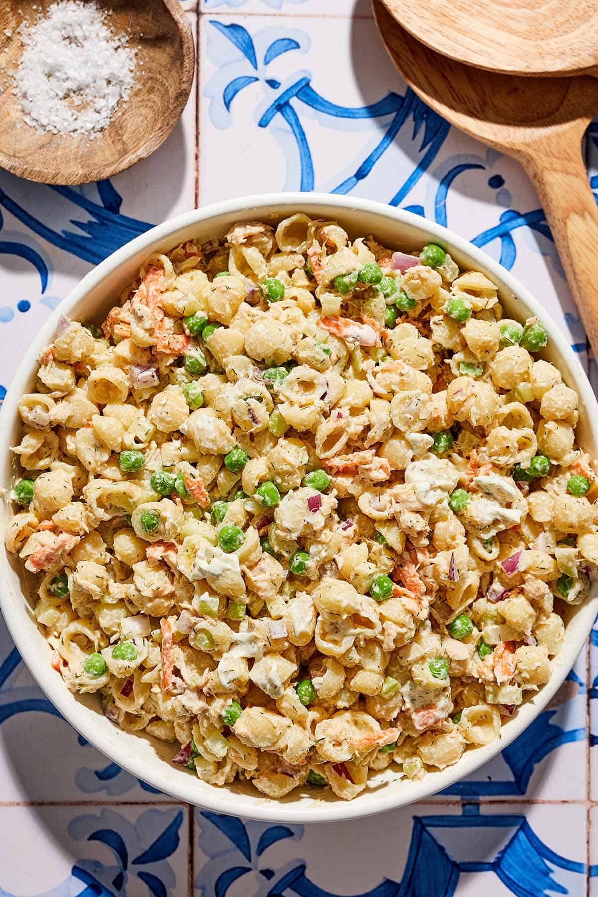 Tuna pasta salad in a serving bowl next to a bowl of salt and wooden serving utensils.