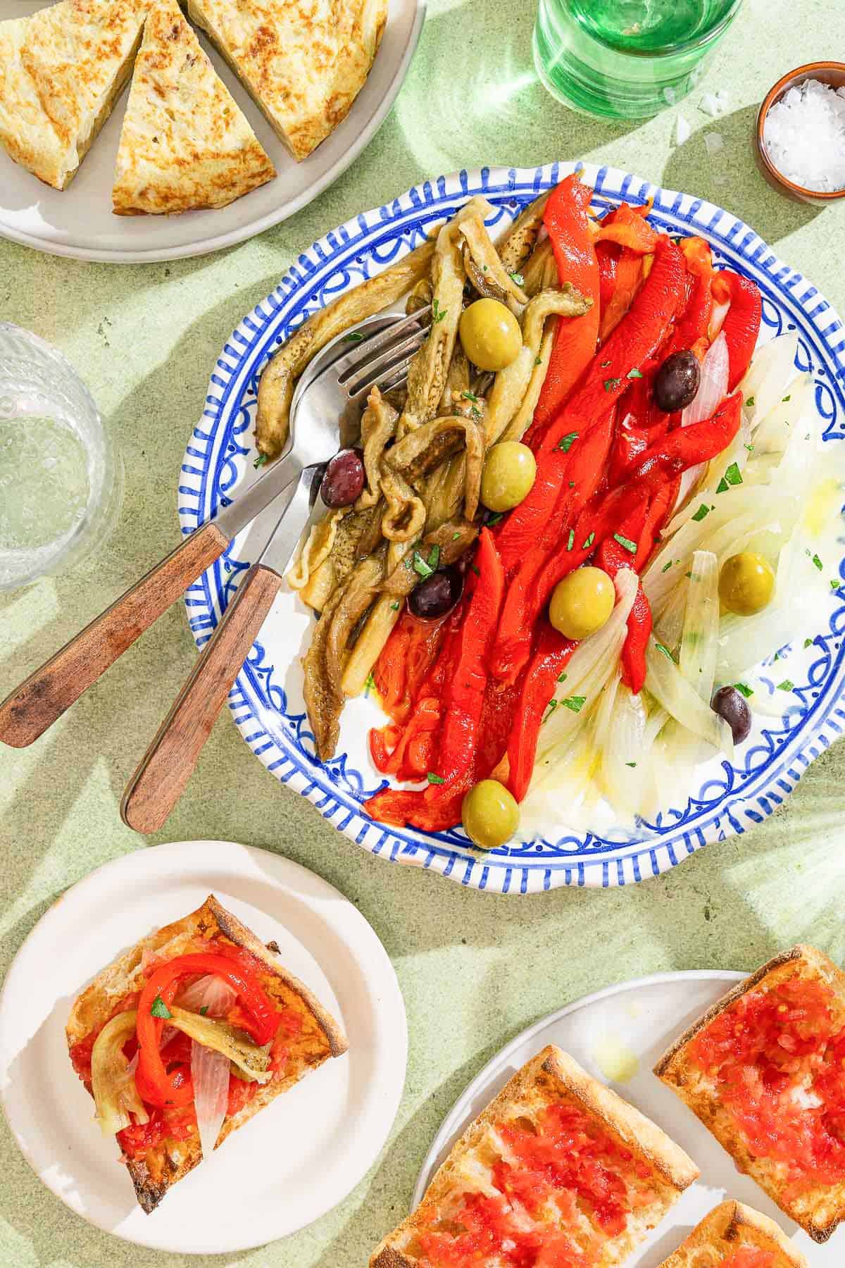Escalivada topped with parsley and olives on a plate with a spoon and a fork. Next to this is a plate with slices of a spanish tortilla, two plates of pan con tomate, a bowl of salt and a bottle of olive oil.