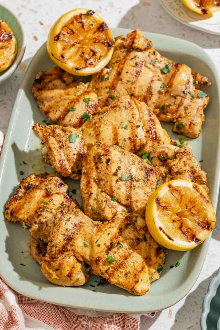Several lemon chicken thighs on a platter with grilled lemon halves.
