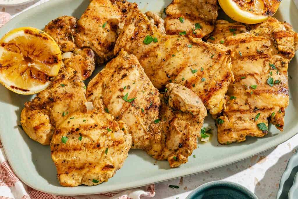 Several lemon chicken thighs on a platter with grilled lemon halves.
