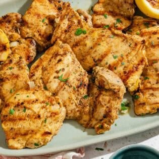 Several lemon chicken thighs on a platter with grilled lemon halves.