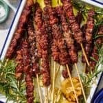 Several arrosticini skewers on a platter with grilled lemon halves and sprigs of rosemary.