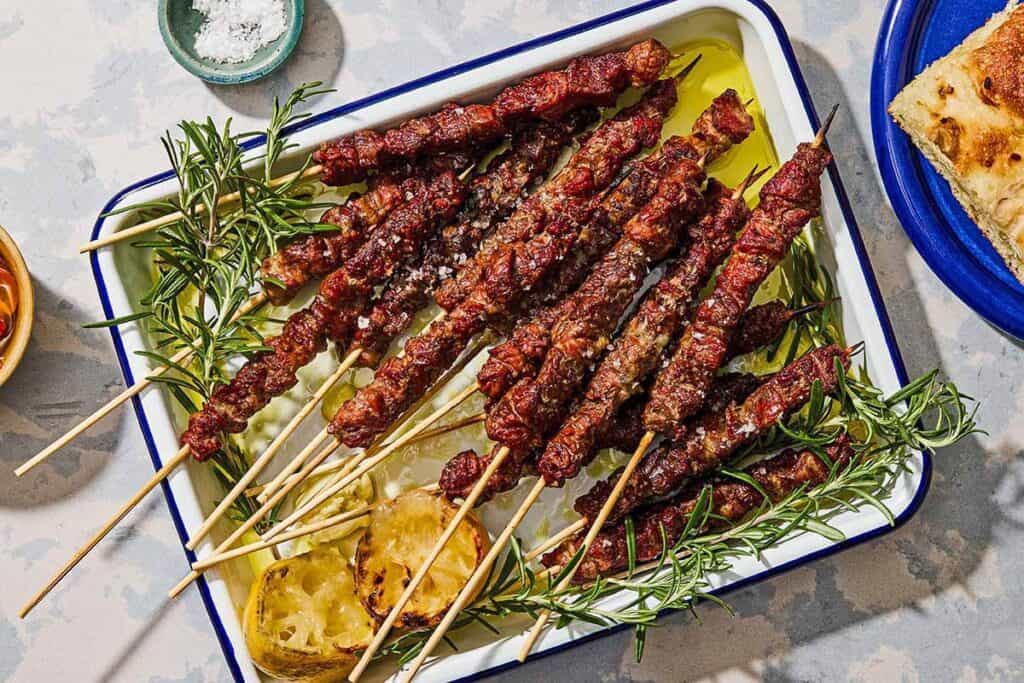 Several arrosticini skewers on a platter with grilled lemon halves and sprigs of rosemary.