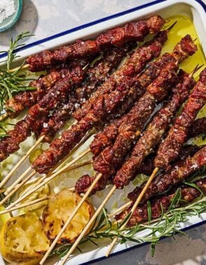 Several arrosticini skewers on a platter with grilled lemon halves and sprigs of rosemary.
