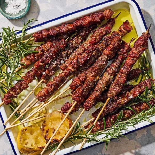 Several arrosticini skewers on a platter with grilled lemon halves and sprigs of rosemary.