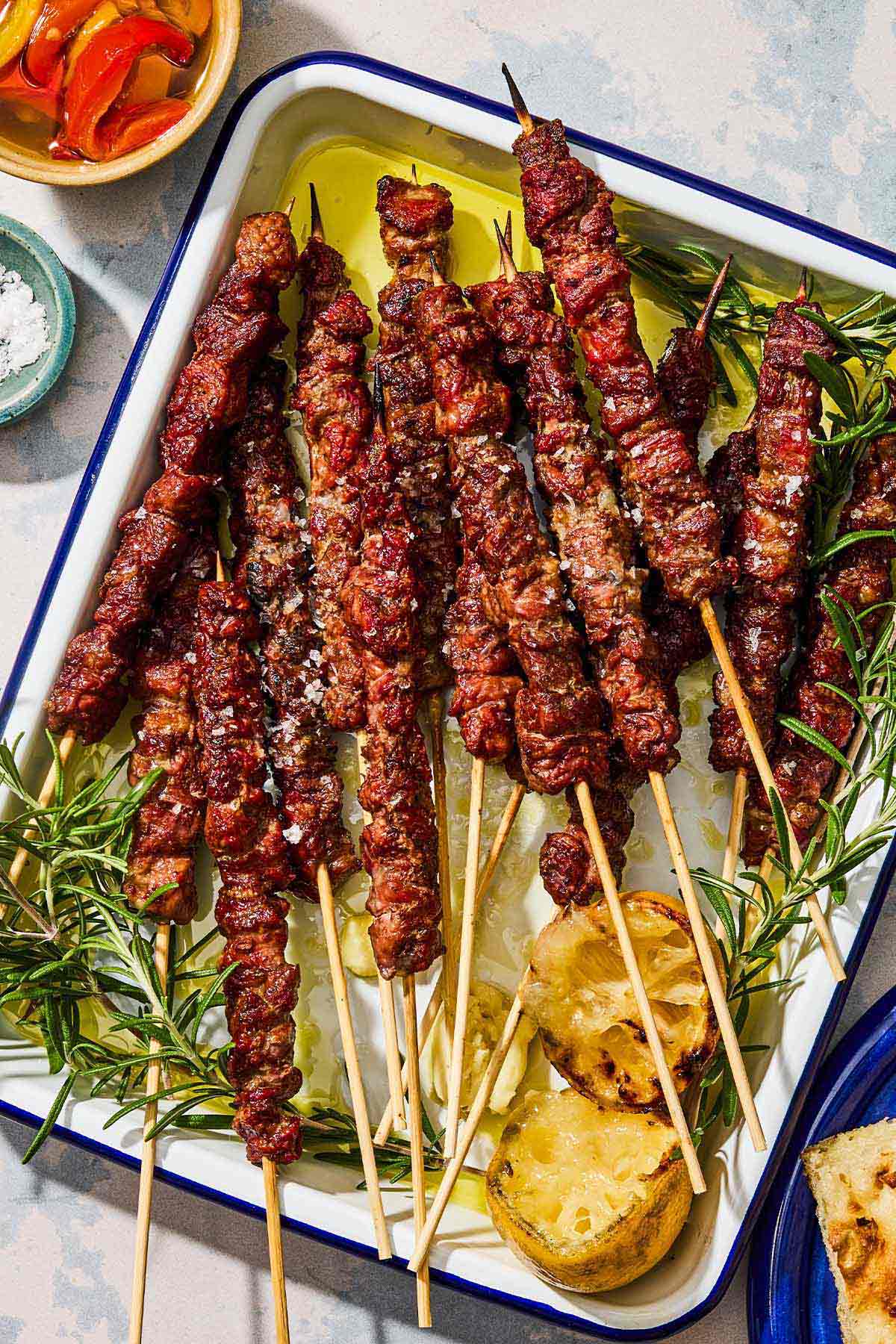 Several arrosticini skewers on a platter with grilled lemon halves and sprigs of rosemary.