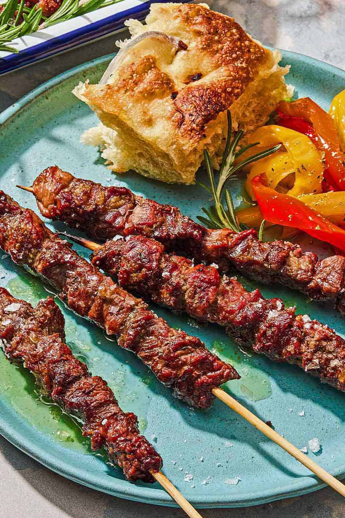 Four arrosticini skewers on a plate with a piece of focaccia and a serving of pepperonata