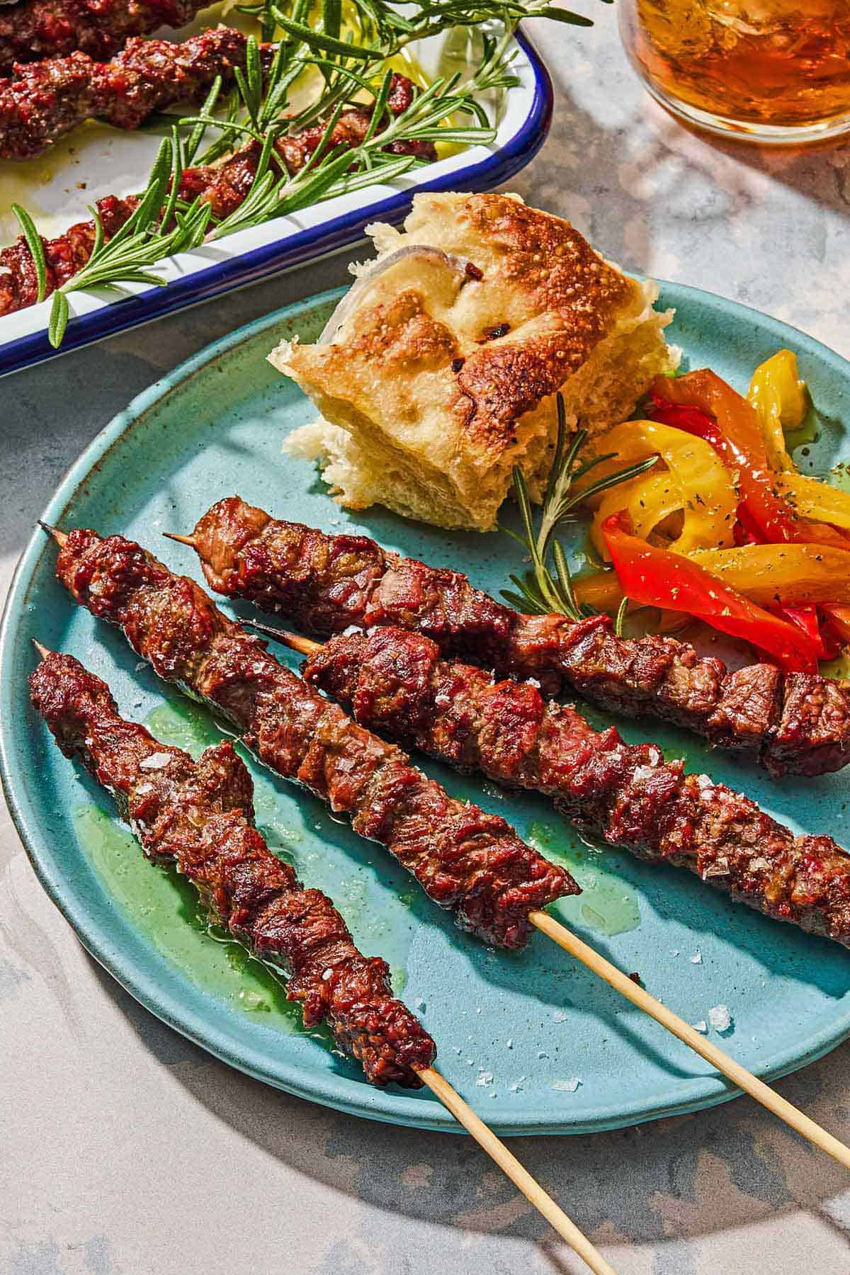 Four arrosticini skewers on a plate with a piece of focaccia and a serving of pepperonata