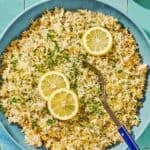 Greek lemon rice topped with lemon wheels in a bowl with a serving spoon next to lemons and a bowl of parsley.