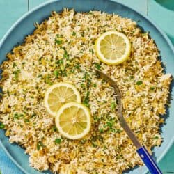 Greek lemon rice topped with lemon wheels in a bowl with a serving spoon next to lemons and a bowl of parsley.