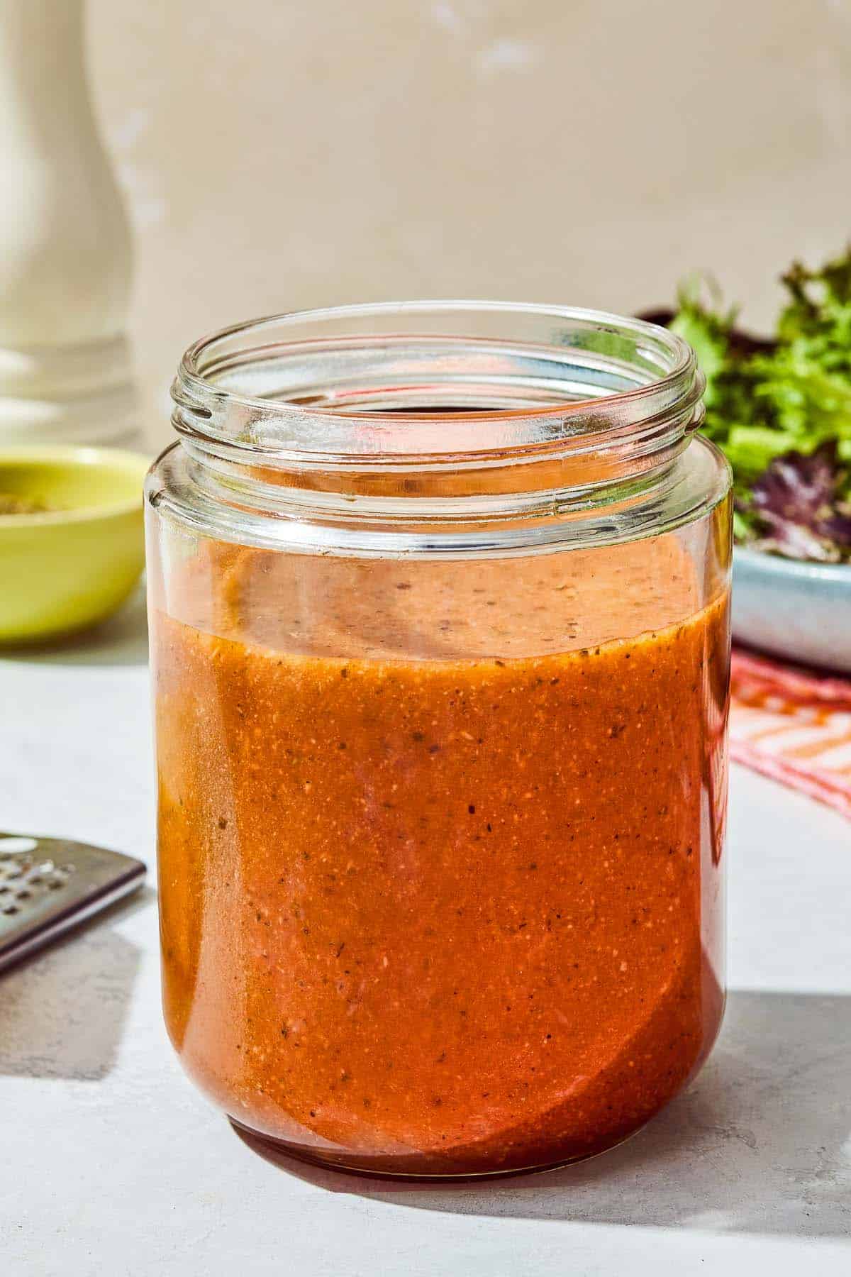 Italian salad dressing in a jar with bowls of lettuce and Italian seasoning in the background.