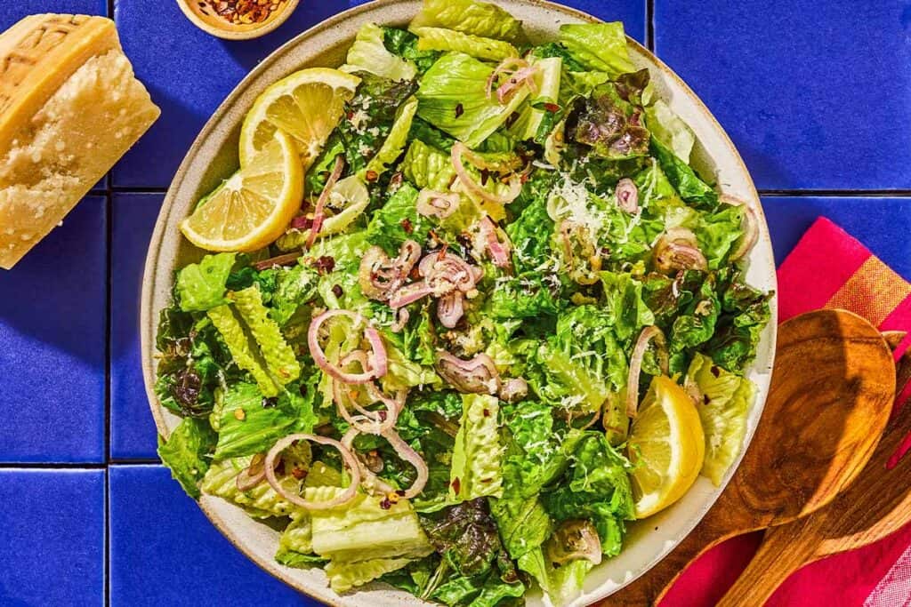 Lemon parmesan lettuce salad in a serving bowl next to a set of wooden serving utensils and a block of parmesan cheese.