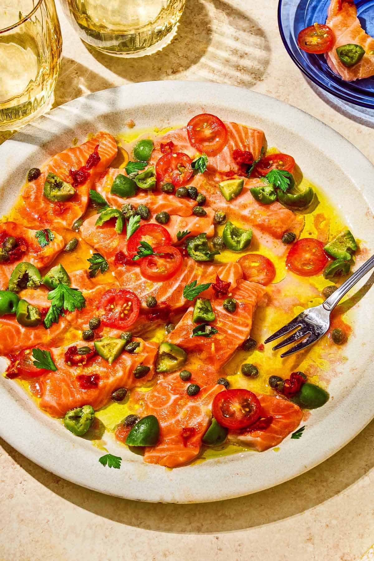 Salmon crudo topped with olives, capers, parsley, chili paste and olive oil on a platter with a fork. Next to this is a serving of the crudo on a plate and 2 glasses of white wine.