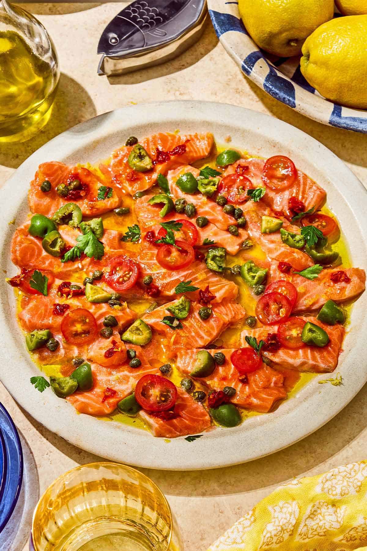 Salmon crudo topped with olives, capers, parsley, chili paste and olive oil on a platter. Next to this is a bowl of lemons, a glass of wine and a bottle of olive oil.