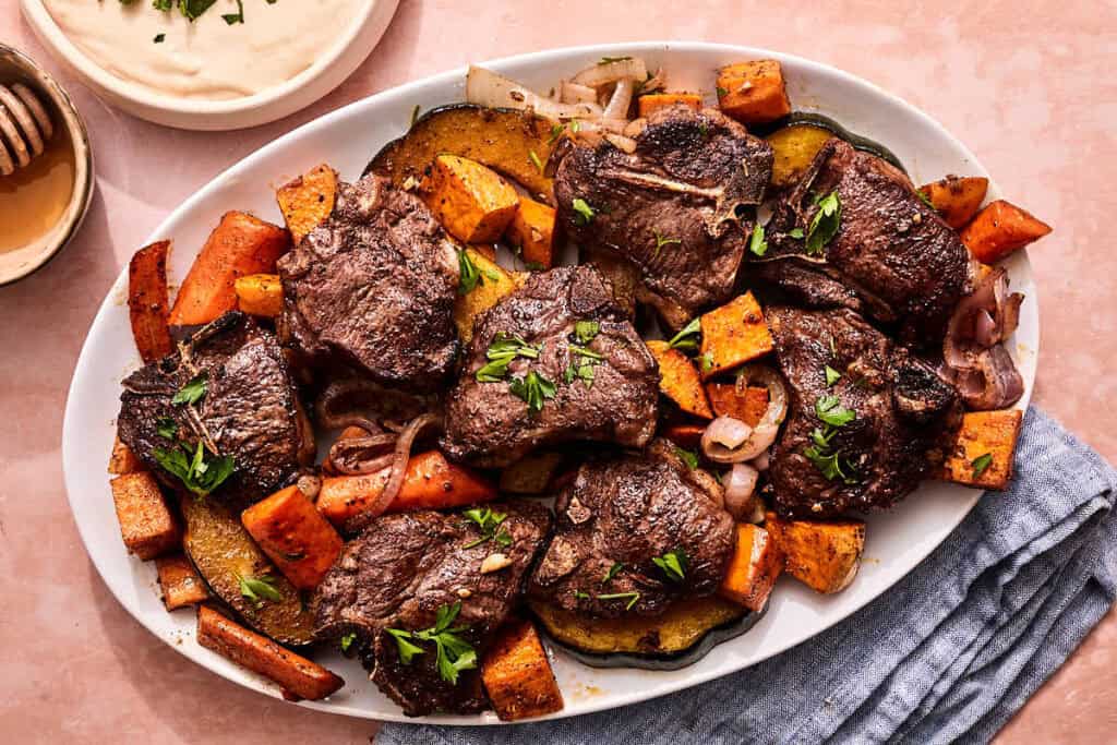 Baked lamb chops with vegetables on a platter next to bowls of tahini sauce and honey.