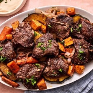 Baked lamb chops with vegetables on a platter next to bowls of tahini sauce and honey.