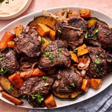 Baked lamb chops with vegetables on a platter next to bowls of tahini sauce and honey.