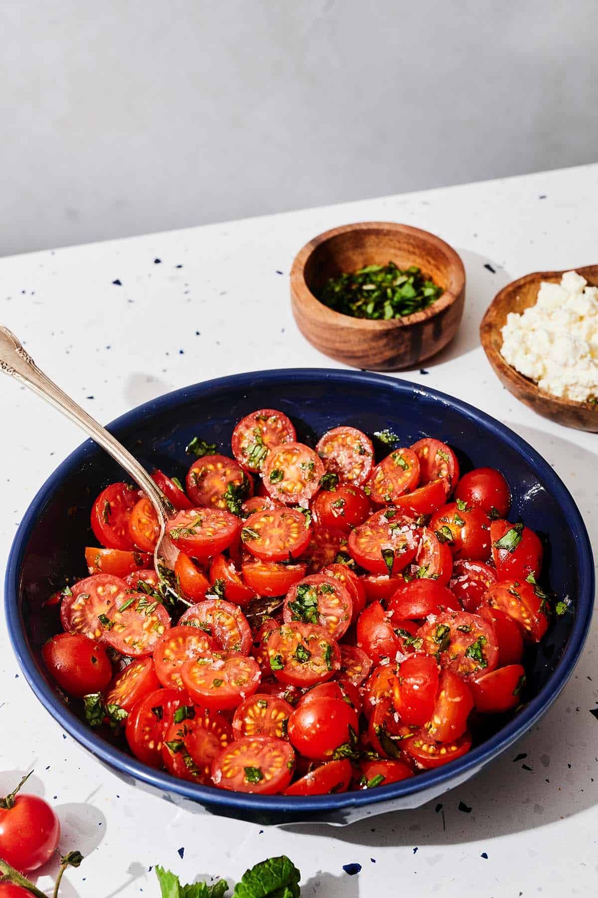 Marinated tomatoes in a bowl with a spoon next to bowls of fresh herbs and feta cheese.