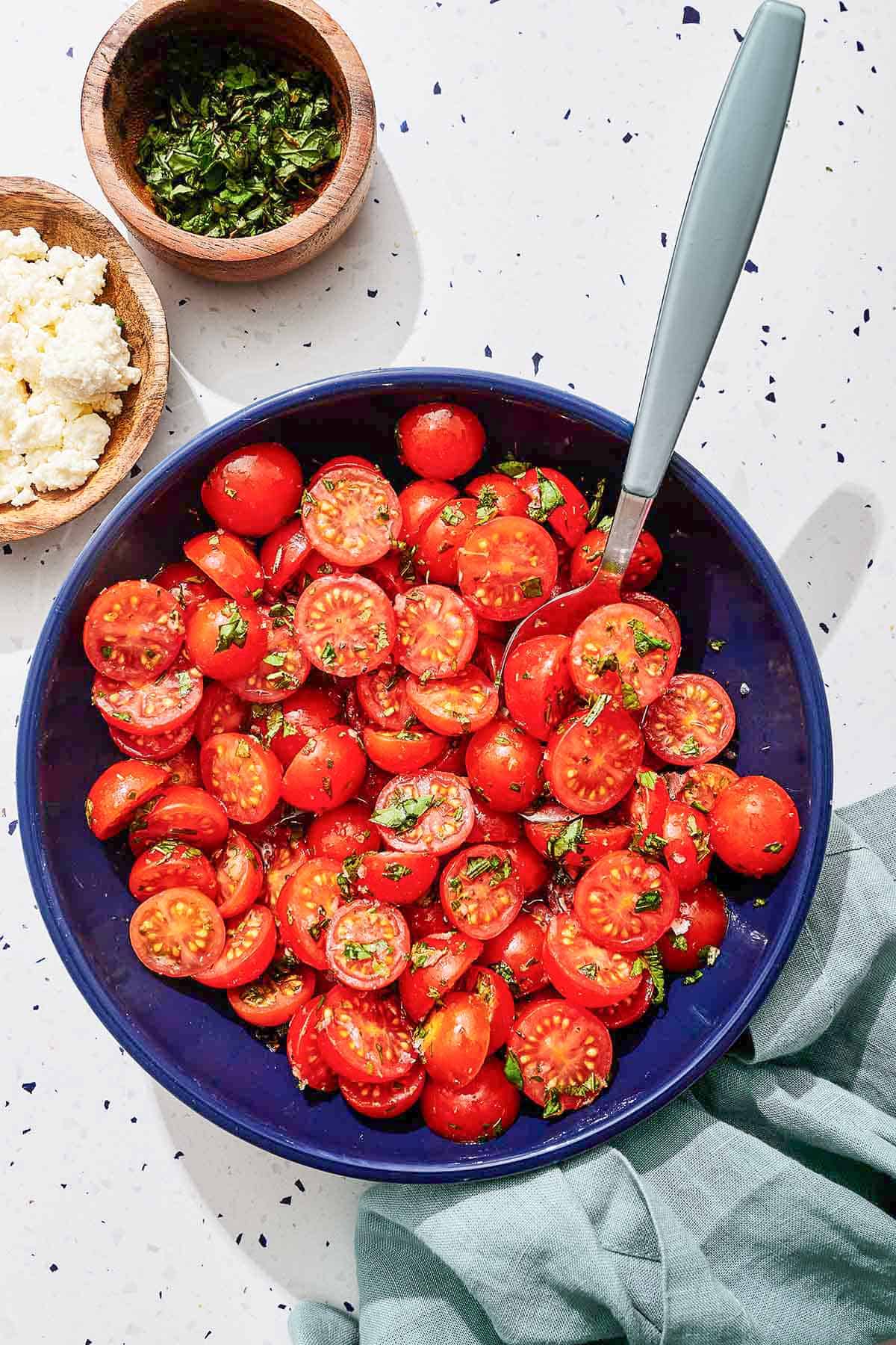 Marinated tomatoes in a bowl with a spoon next to bowls of fresh herbs and feta cheese.