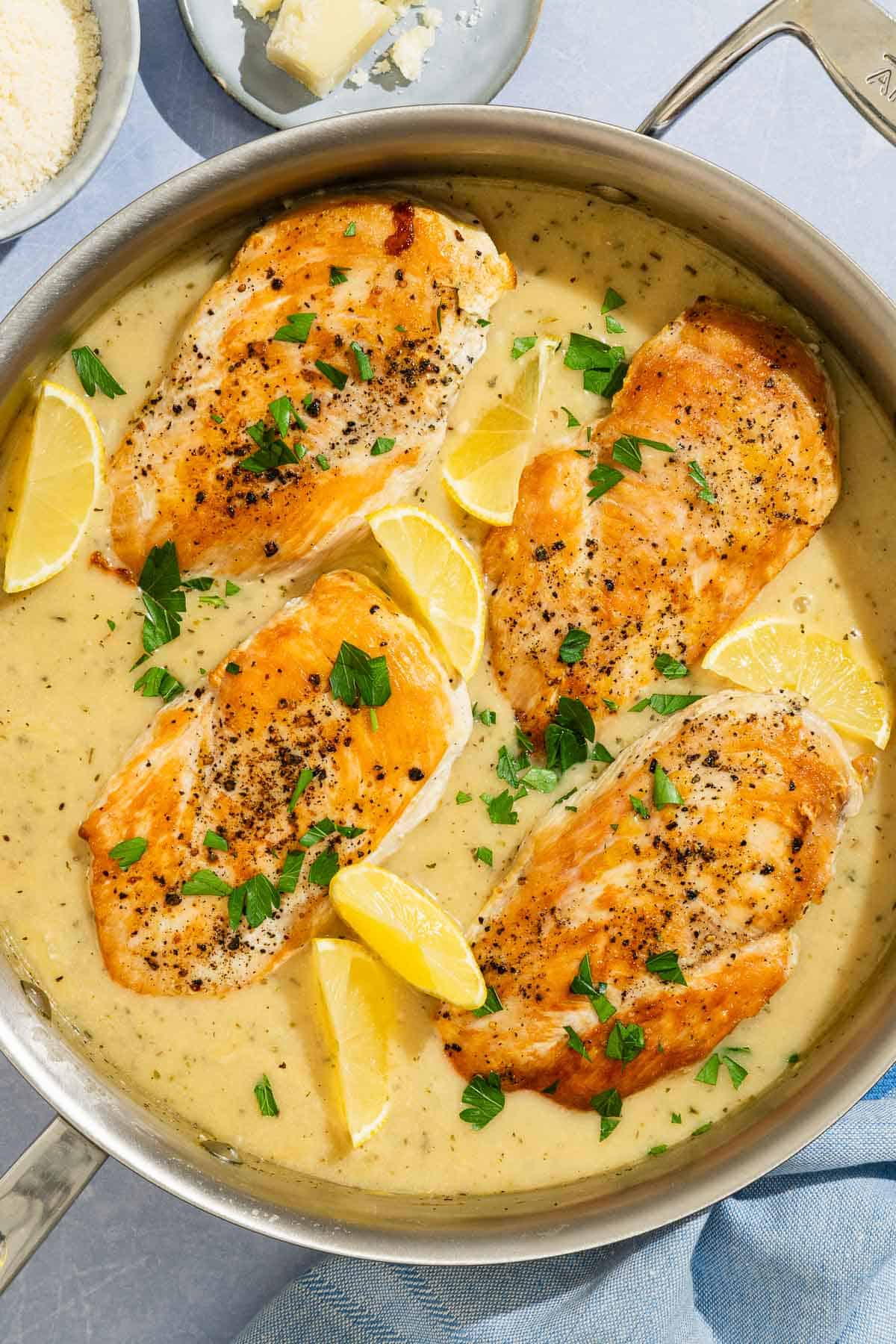 Four garlic parmesan chicken fillets topped with sauce and parsley in a skillet with lemon wedges. Next to this is a bowl of grated Parmigiano-Reggiano cheese along with a small plate with a cheese wedge.