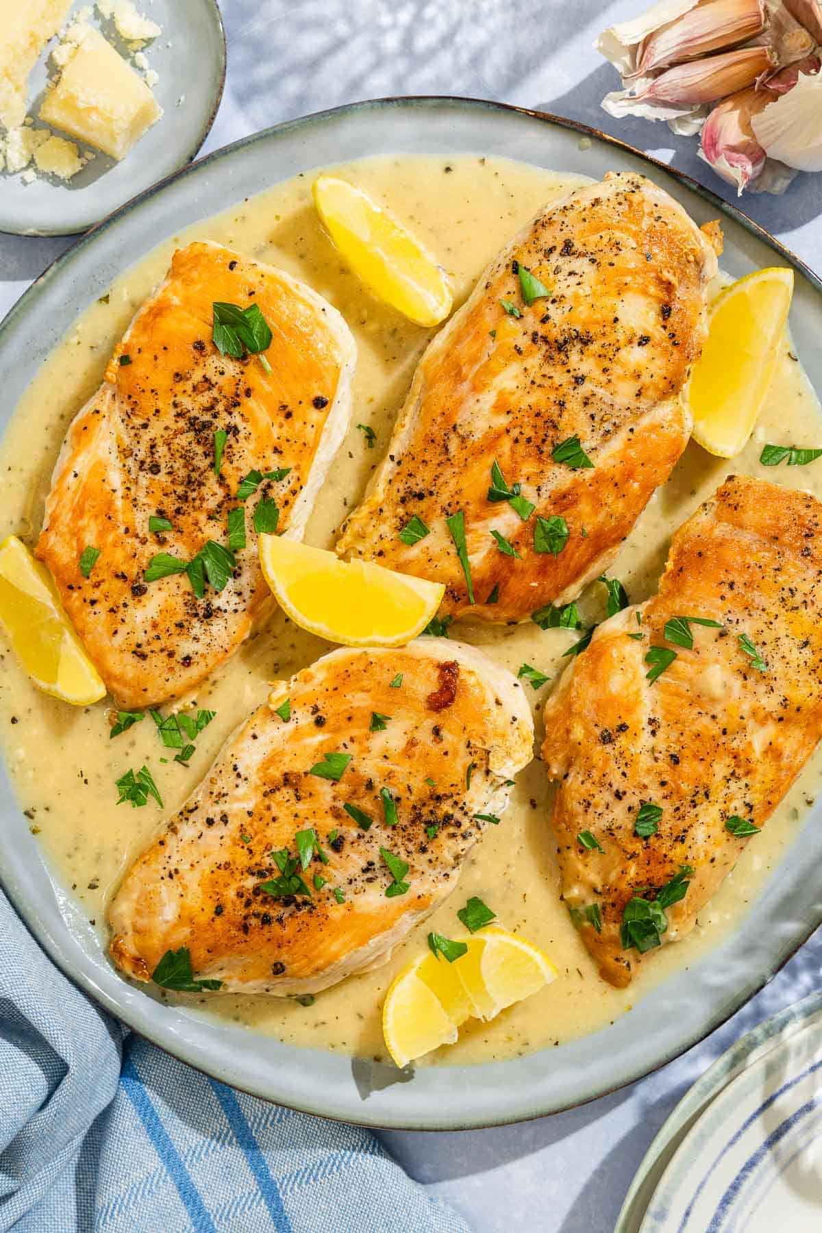 Four garlic parmesan chicken fillets topped with sauce and parsley on a plate with lemon wedges. Next to this is a plate with a cheese wedge and several cloves of garlic.